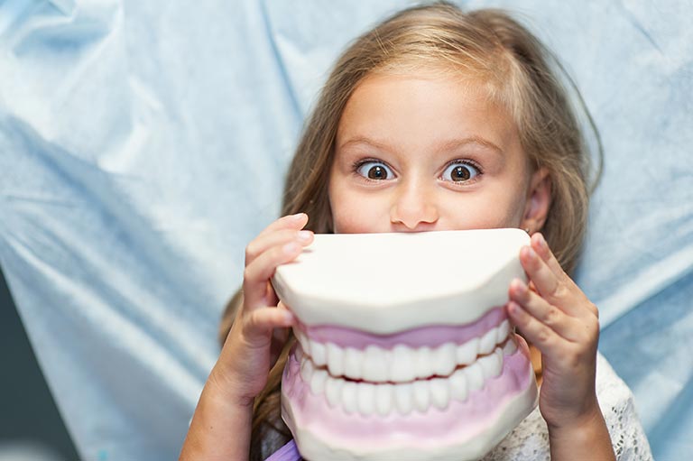 A little girl humorously holds up a massive set of dentures to her face.